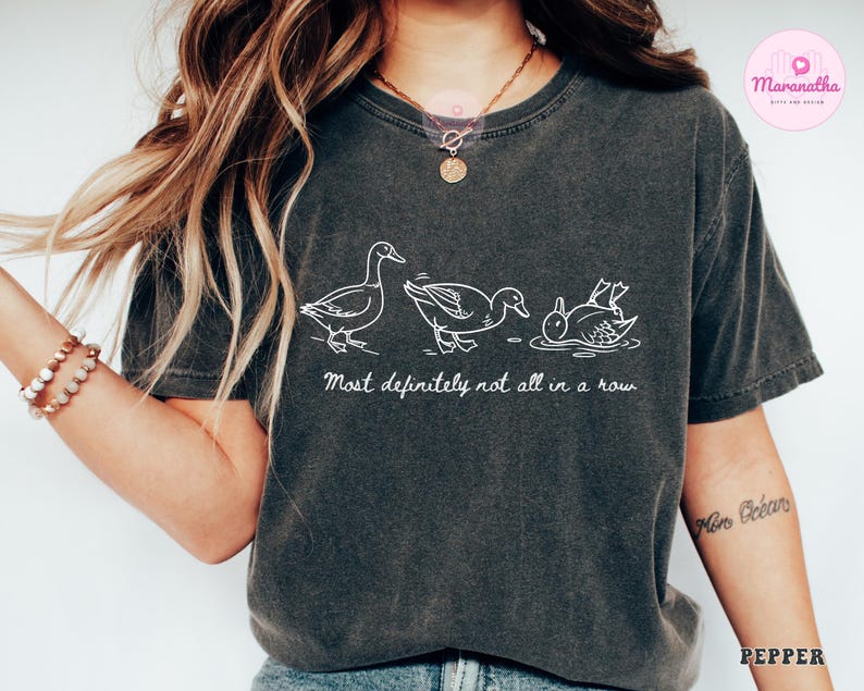 May include: Dark gray t-shirt with a white line drawing of three ducks, one upside down. The text "Most definitely not all in a row" is below the ducks. The shirt is a casual style.