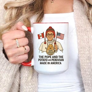 May include: White ceramic mug with a red interior and handle. The mug features a cartoon illustration of a figure holding Peruvian and American flags, with the text "THE POPE AND THE POTATO ARE PERUVIAN MADE IN AMERICA."