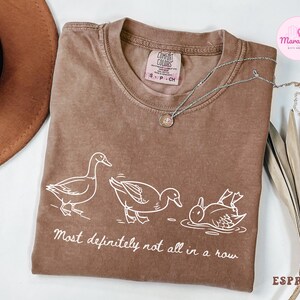 May include: Espresso-colored t-shirt with a white graphic of three ducks in different poses, with the text "Most definitely not all in a row." A silver necklace with a pendant rests on the shirt. A brown hat is visible in the upper left corner.