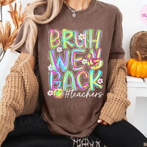 K&ouml;nnte beinhalten: Ein braunes T-Shirt mit dem Schriftzug "BRUH WE BACK" in bunten, umrandeten Buchstaben, verziert mit kleinen Blumen. Der Hashtag "#teachers" befindet sich darunter. Das T-Shirt geh&ouml;rt zu einer Kollektion zum Thema Lehrer.