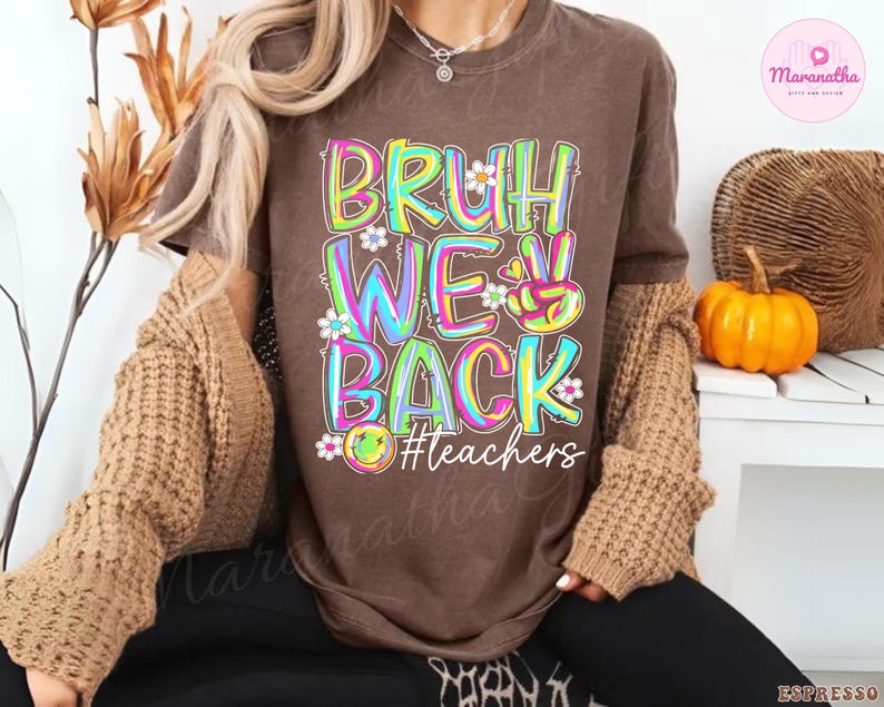 K&ouml;nnte beinhalten: Ein braunes T-Shirt mit dem Schriftzug "BRUH WE BACK" in bunten, umrandeten Buchstaben, verziert mit kleinen Blumen. Der Hashtag "#teachers" befindet sich darunter. Das T-Shirt geh&ouml;rt zu einer Kollektion zum Thema Lehrer.