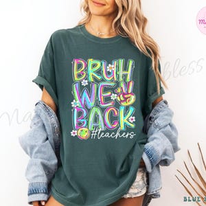 K&ouml;nnte beinhalten: Dunkelgr&uuml;nes T-Shirt mit dem Text "BRUH WE BACK" in bunten, umrandeten Buchstaben. Das Design beinhaltet ein Friedenszeichen, Blumen und ein Smiley-Gesicht, mit dem Hashtag "#teachers" darunter. Das Shirt hat kurze &Auml;rmel.