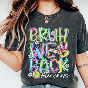 K&ouml;nnte beinhalten: Dunkelgraues T-Shirt mit dem Schriftzug "BRUH WE BACK" in bunten Buchstaben. Das Design beinhaltet ein Peace-Zeichen, G&auml;nsebl&uuml;mchen und ein Smiley. Der Hashtag "#teachers" ist ebenfalls im Design enthalten.