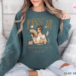 Puede incluir: Una sudadera azul turquesa con un dise&ntilde;o gr&aacute;fico que presenta a una mujer sosteniendo un abanico y un vaso de t&eacute; helado. El texto en la sudadera dice "Where them fans at Hot Flashes and Iced Tea Social Club".