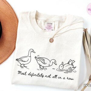 May include: Ivory-colored sweatshirt with a black line drawing of three ducks, one standing, one bending, and one in the water. The text "Most definitely not all in a row" is below the ducks. A gold necklace is on the shirt.