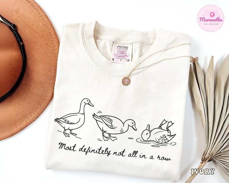 May include: Ivory-colored sweatshirt with a black line drawing of three ducks, one standing, one bending, and one in the water. The text "Most definitely not all in a row" is below the ducks. A gold necklace is on the shirt.
