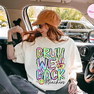 K&ouml;nnte beinhalten: Cremefarbenes Sweatshirt mit dem Schriftzug "BRUH WE BACK" in bunten Buchstaben. Das Design beinhaltet ein Peace-Zeichen, Blumen und ein Smiley. Der Hashtag "#teachers" ist ebenfalls enthalten.