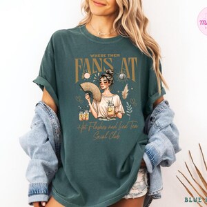Puede incluir: Una camiseta azul turquesa con un dise&ntilde;o gr&aacute;fico que presenta a una mujer con un abanico, el texto "Where Them Fans At" y las palabras "Hot Flashes and Iced Tea Social Club".