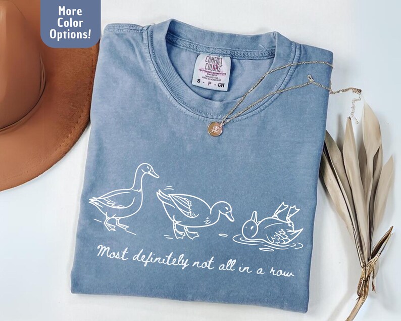 May include: A blue Comfort Colors t-shirt with a white graphic of three ducks in different poses and the text "Most definitely not all in a row." A gold necklace is draped across the shirt.