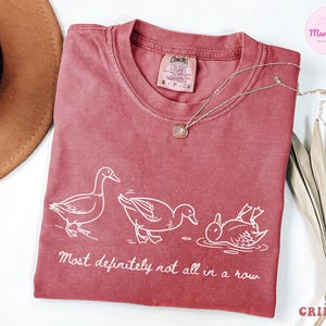 May include: Crimson-colored t-shirt with a white graphic of three ducks in different poses, with the text "Most definitely not all in a row." A gold necklace and a brown hat are also visible.