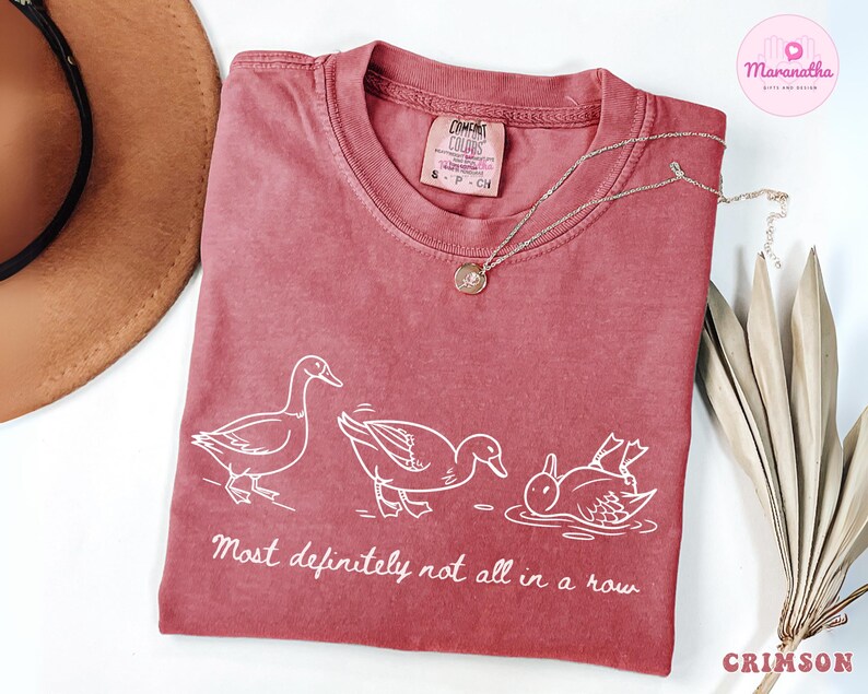 May include: Crimson-colored t-shirt with a white graphic of three ducks in different poses, with the text "Most definitely not all in a row." A gold necklace and a brown hat are also visible.