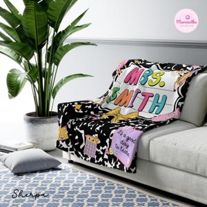 May include: A black and white Sherpa blanket with the name "MRS. SMITH" in colorful letters. The blanket has a school theme with images of a calculator, ABCs, and a bow. The blanket is draped over a white couch.