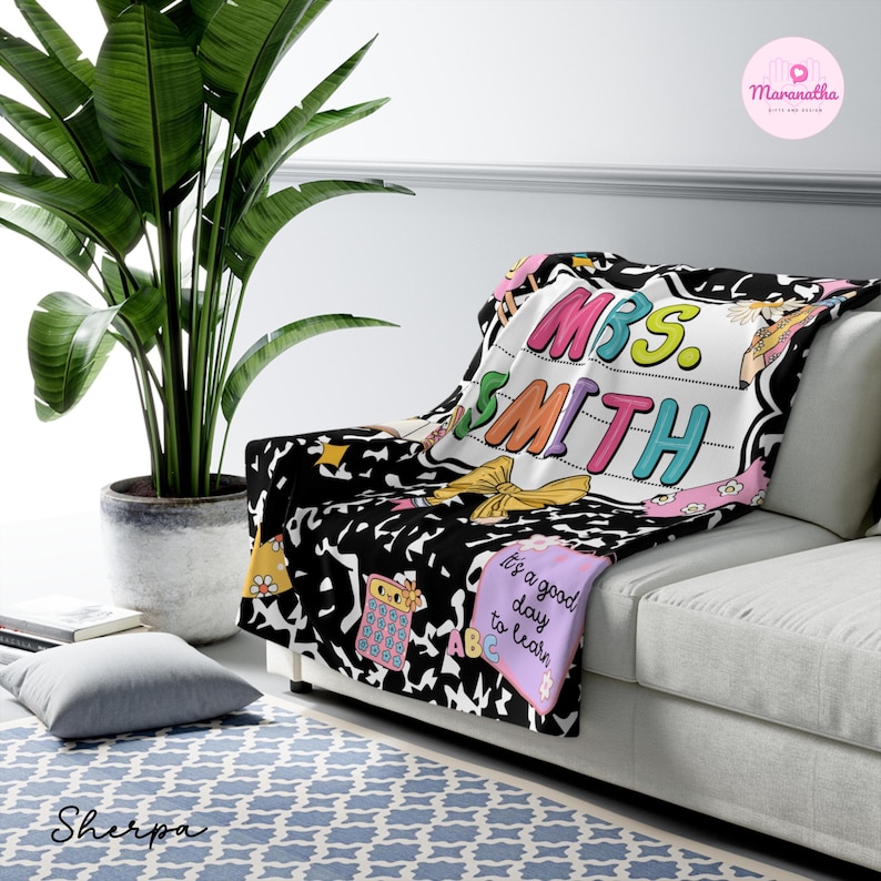 May include: A black and white Sherpa blanket with the name "MRS. SMITH" in colorful letters. The blanket has a school theme with images of a calculator, ABCs, and a bow. The blanket is draped over a white couch.