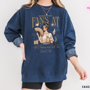Puede incluir: Una sudadera azul marino con un gr&aacute;fico de una mujer sosteniendo un abanico y un vaso de t&eacute; helado. El texto "Where Them Fans At" est&aacute; por encima del gr&aacute;fico, y el texto "Hot Flashes and Iced Tea Social Club" est&aacute; por debajo del gr&aacute;fico.