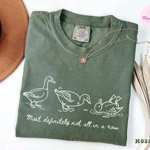 May include: Moss green t-shirt with a white graphic of three ducks, one standing, one mid-motion, and one upside down in water. The text "Most definitely not all in a row" is below the ducks.