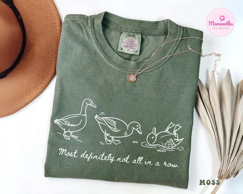 May include: Moss green t-shirt with a white graphic of three ducks, one standing, one mid-motion, and one upside down in water. The text "Most definitely not all in a row" is below the ducks.