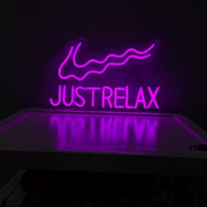 Puede incluir: Un letrero de neón morado con una línea ondulada y el texto "JUST RELAX".