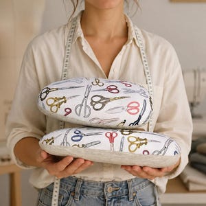 May include: Two oval-shaped sewing tailor hams featuring a white fabric top printed with a colorful pattern of various scissors in shades of gold, red, blue, and black, paired with a light beige textured fabric base for pressing and shaping seams.