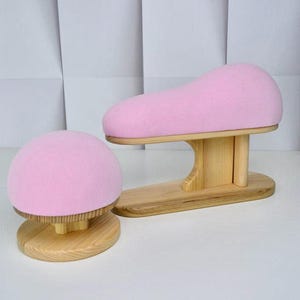 Può includere: Due forme per cappelli in legno. Una è a forma di cupola arrotondata, l'altra è curva. Entrambe hanno una parte superiore imbottita rosa chiaro e morbida. Le basi sono in legno chiaro.