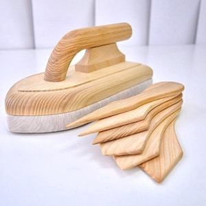 Può includere: Un ferro da stiro in legno con manico curvo e una pila di utensili in legno per la modellatura. Il ferro ha una finitura a venatura chiara e una base in tessuto bianco. Anche gli utensili sono in legno chiaro.