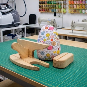 Può includere: Un battente da sarto in legno, un tampone da stiro e un asse per maniche su un tappetino da taglio verde. Un tampone da stiro a forma di uovo colorato con un motivo a bottoni. Materiali e attrezzature per cucire sullo sfondo.