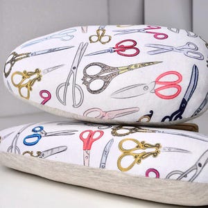 May include: Two oblong-shaped decorative pillows with a white background and a pattern of various scissors in different colors and styles. The scissors are in shades of gold, pink, blue, and black. The pillows are stacked on top of each other.
