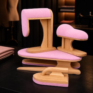 May include: A set of four wooden tailoring tools with light pink fabric padding. The collection includes a tall vertical press, a curved sleeve board, a flat ironing board, and a wooden clapper with a handle, all featuring natural wood grain.