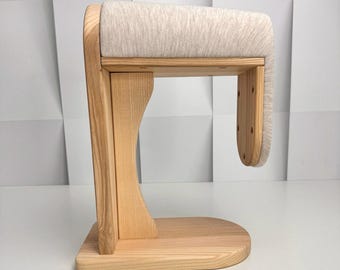 May include: A wooden jewelry display stand with a light beige padded top. The stand is made of light-colored wood and has a curved design. The base is rectangular with rounded edges. The padded top is approximately 8 inches long.