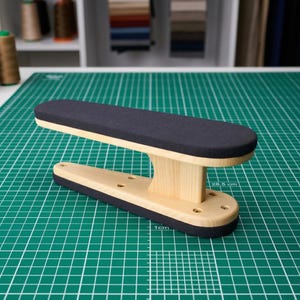 May include: A wooden tailor's clapper with a dark gray padded top and bottom. The clapper is made of light-colored wood and has a curved design. The clapper is 28.5 cm long and 1 cm wide.