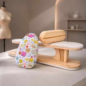 May include: A wooden tailor's ham and sleeve board set. The tailor's ham is covered in white fabric featuring a colourful pattern of various buttons in shades of pink, yellow, blue, and orange. The sleeve board is made of light-coloured natural wood.