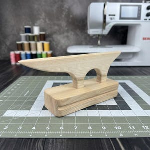 Può includere: Un clapper di legno chiaro, uno strumento per cucire, con una base rettangolare e una parte superiore curva. Il clapper è posizionato su un tappetino da taglio grigio con una macchina da cucire e rocchetti di filo sullo sfondo.