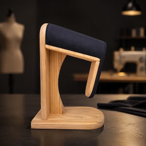 May include: A wooden tailoring ham press with a curved, light-toned wood base and vertical support. The top section is covered in a smooth, dark navy blue fabric, designed for pressing curved seams and garment details during the sewing process.