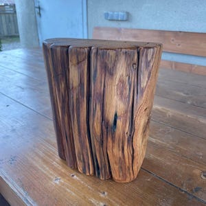 Pode incluir: Um bloco de madeira r&uacute;stico com um acabamento natural e desgastado. O bloco tem uma forma retangular com bordas arredondadas e uma superf&iacute;cie &aacute;spera e texturizada. A madeira &eacute; de uma cor castanho escura com veios de castanho escuro e preto.
