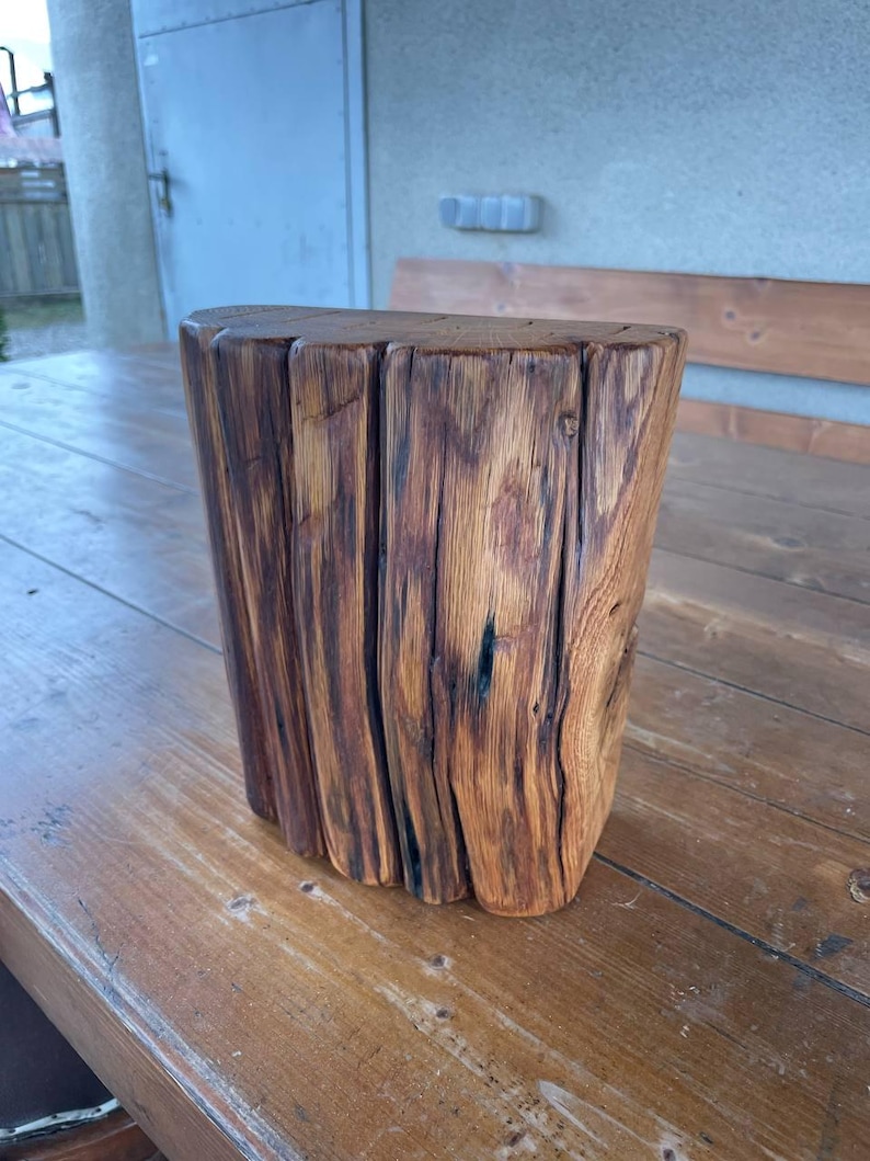 Pode incluir: Um bloco de madeira r&uacute;stico com um acabamento natural e desgastado. O bloco tem uma forma retangular com bordas arredondadas e uma superf&iacute;cie &aacute;spera e texturizada. A madeira &eacute; de uma cor castanho escura com veios de castanho escuro e preto.