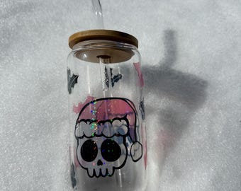 May include: A clear glass tumbler with a wooden lid and a straw. The tumbler has a pink and white design of a skull wearing a Santa hat and holly leaves.