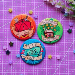 Puede incluir: Tres pines redondos y coloridos con temas de Dungeons & Dragons. Un pin presenta un dado rojo con el texto "WELL, SHIT". Otro tiene un fondo verde con el texto "DUNGEON MASTER". El tercer pin es azul con el texto "DICE DON'T LIE".