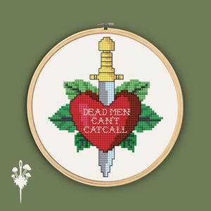May include: Cross-stitch embroidery in a wooden hoop. The design features a red heart pierced by a sword, surrounded by green leaves. The text "DEAD MEN CAN'T CATCALL" is stitched in white. The background is white.