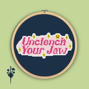 May include: A cross-stitch embroidery hoop with the phrase "Unclench Your Jaw" in pink letters on a navy blue background. The design includes yellow star accents and a smiling face. The hoop is set against a light green backdrop.