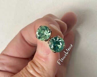 May include: Two round, light green stud earrings with a faceted design. The earrings are set in silver-colored metal. The image includes the text "Fleurbleue".
