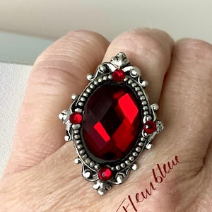 May include: A silver ring with a large red gemstone surrounded by smaller red gemstones. The ring has a detailed, ornate design.