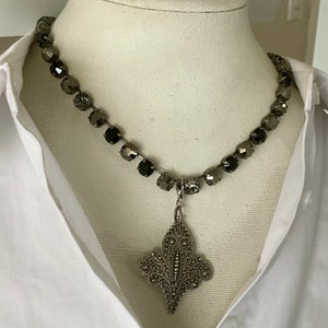 Puede incluir: Un collar con un colgante plateado en forma de flor de lis. El collar presenta cuentas facetadas de color gris oscuro. El colgante está intrincadamente detallado con un diseño floral. El collar se muestra sobre una camisa blanca.