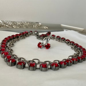 Op de afbeelding: Een zilverkleurige ketting en bijpassende oorbellen set. De sieraden hebben een reeks levendige rode edelstenen gezet in een zilveren metalen ketting. De oorbellen hebben een enkele rode edelsteen. De sieraden worden op een wit oppervlak getoond.