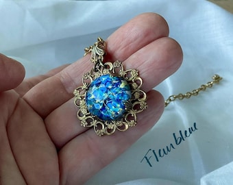 Könnte beinhalten: Eine goldene Halskette mit einem blauen und grünen, schillernden Glasanhänger. Der Anhänger ist von einem goldenen Filigranmuster umgeben.