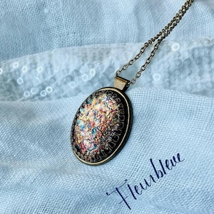 Puede incluir: Un collar de cadena de bronce con un colgante ovalado que presenta un diseño de vidrio iridiscente y colorido. El colgante tiene un borde texturizado y metálico. El texto "Fleurbleue" es visible en la esquina inferior derecha de la imagen.
