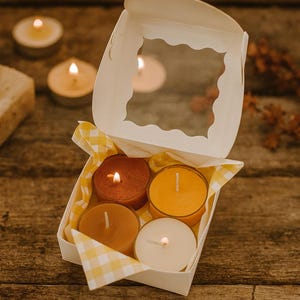 May include: A white cardboard gift box with a scalloped window lid containing four small round candles in shades of red, orange, tan, and white. The box is lined with yellow and white chequered tissue paper, and each candle has a lit wick.