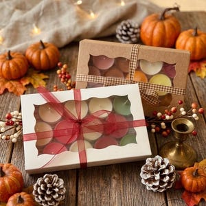May include: Two rectangular gift boxes with clear windows containing assorted tea light candles. The white box is tied with a red ribbon, and the brown box is tied with a checkered ribbon. Both boxes are surrounded by small pumpkins and pinecones.