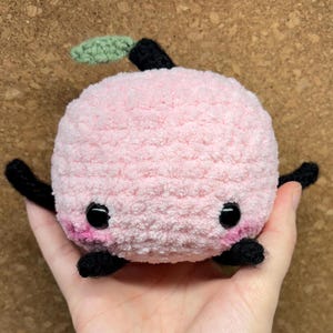May include: A pink crocheted peach plush toy with black eyes and a green leaf stem. The peach has a fuzzy texture and black crocheted arms and legs.
