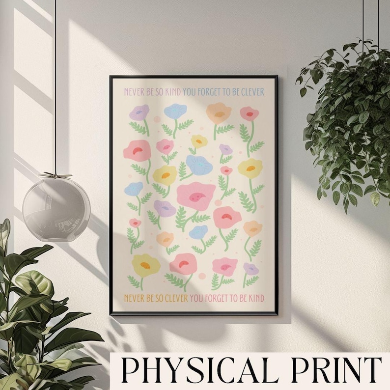 Bright neutral room with soft window light and a leafy plant in the corner, featuring a framed floral artwork on the wall. The design shows scattered pastel flowers in pink, yellow, blue, and purple with soft green stems on a light background. Text at the top reads “NEVER BE SO KIND YOU FORGET TO BE CLEVER” and at the bottom “NEVER BE SO CLEVER YOU FORGET TO BE KIND,” making it perfect for soft, feminine or cottage-inspired decor