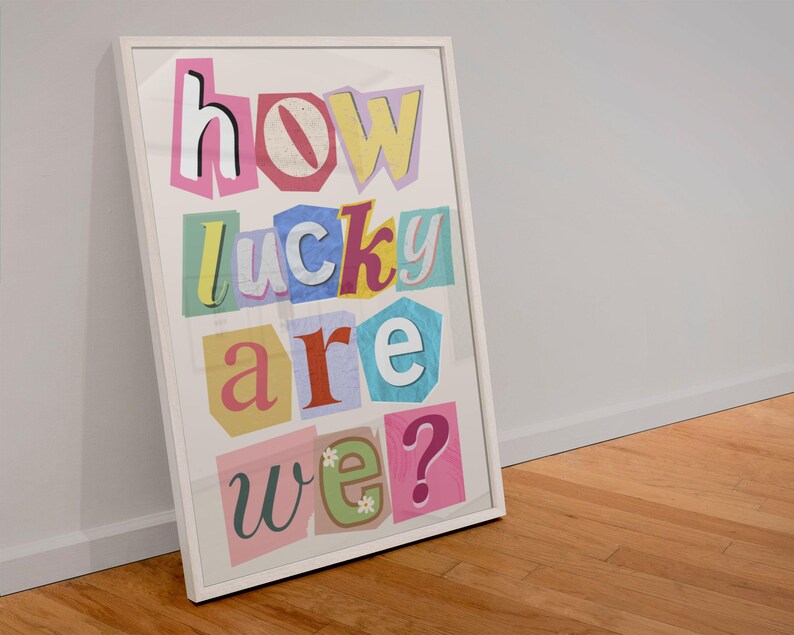 May include: A framed art print with the words "how lucky are we?" in a colorful, cut-out style. The letters are in various colors, including pink, yellow, blue, and red, set against a white background. The frame is white.