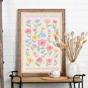 Minimal interior with a light wall, wooden console table, and decorative dried stems, showcasing a framed floral artwork. The design features scattered pastel flowers with soft green stems and text reading “NEVER BE SO KIND YOU FORGET TO BE CLEVER” and “NEVER BE SO CLEVER YOU FORGET TO BE KIND,” creating a gentle, uplifting piece that suits feminine, cozy, or cottagecore-inspired interiors